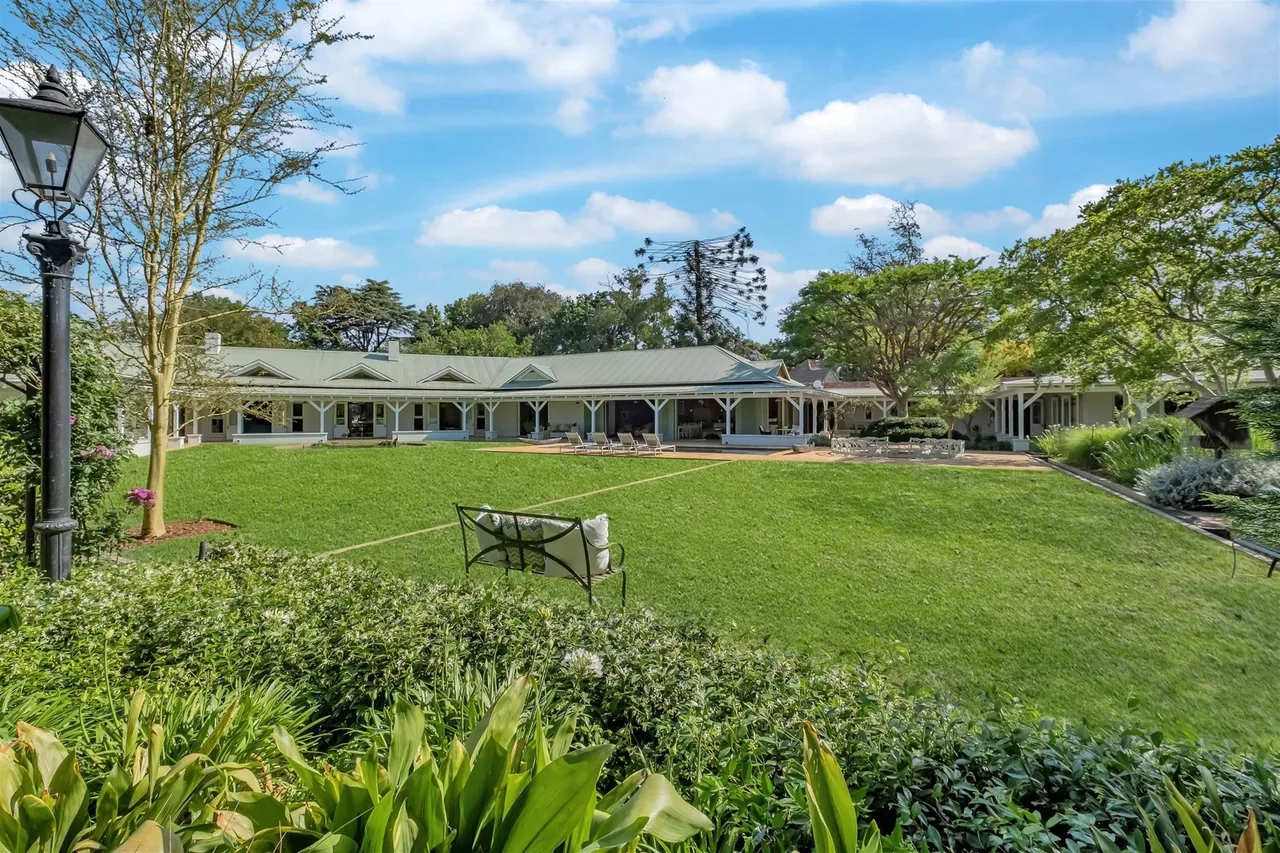 A long, white single-storey Melrose home with a green roof and a deep columned veranda, behind a wide lawn framed by mature trees, a garden bench and a lamp post