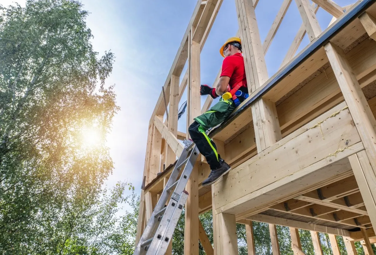 A construction worker in a hard hat and red shirt sitting on the timber frame of a new house, with a ladder beside him and sun through the trees