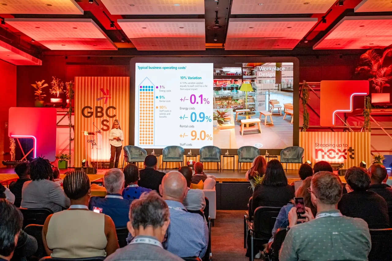 A speaker presents on a stage at a GBCSA Green Building Convention, with a slide on typical business operating costs and a seated audience in the foreground under red lighting